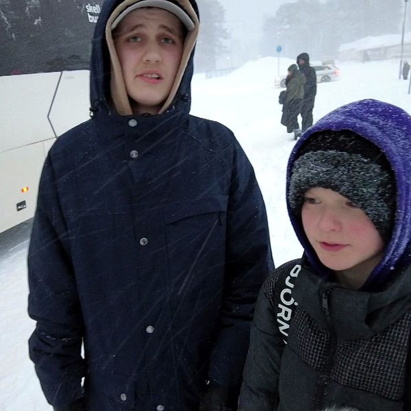 Skolbarnen Alvar och Liam  står framför en vit buss med huvorna uppdragna. Det snöar kraftigt.