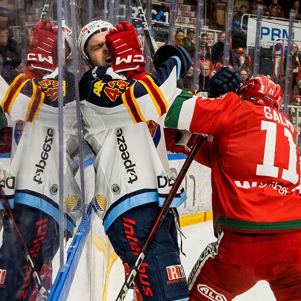 Två hockeyspelare kämpar om pucken vid rinken. Reflektion i glaset.