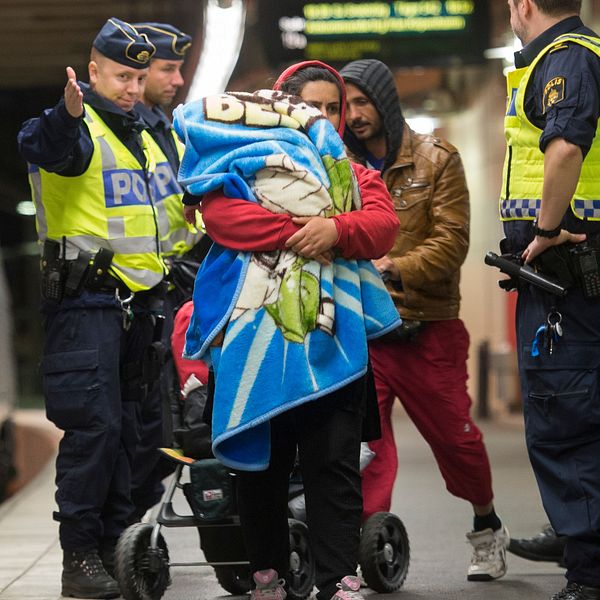 Poliser interagerar med en person som håller ett barn insvept i en filt på en tågstation, medan en andra polis övervakar situationen. I bakgrunden står ett tåg stilla på perrongen.