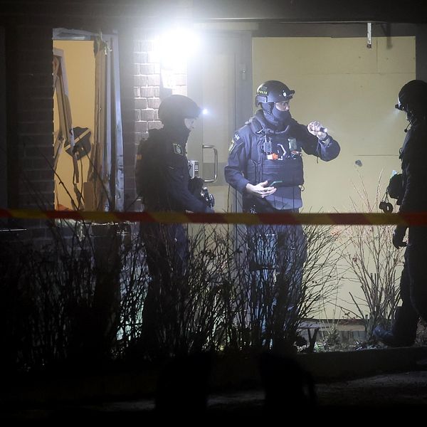 Poliser iklädda taktisk utrustning står utanför ett upplyst rum, med avspärrningsband i förgrunden, medan de genomför en insats på natten.