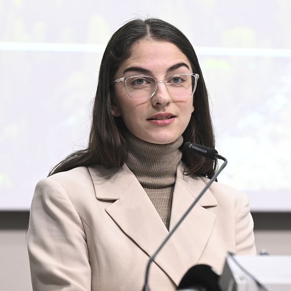 Kvinna i beige kavaj vid ett podium, talar vid en konferens.