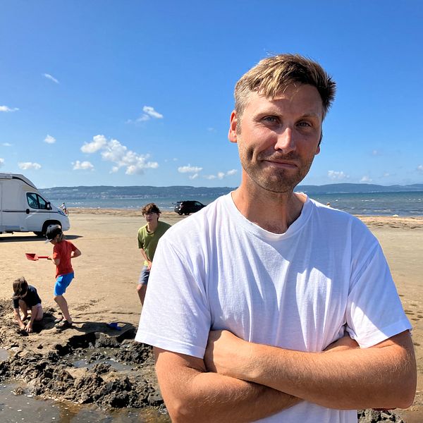 Pappan Daniel Norling på stranden i Laholm med lekande barn i bakgrunden.