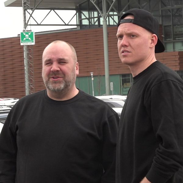 Två personer, män, iklädda svarta tröjor står på en parkeringsplats.