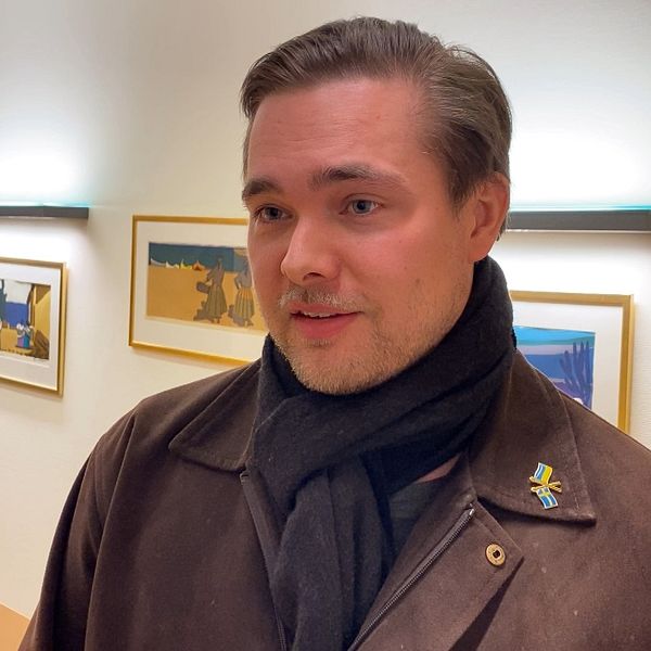 Man i brun jacka och mörk halsduk i ett kontorsrum med tavlor på väggen i bakgrunden.