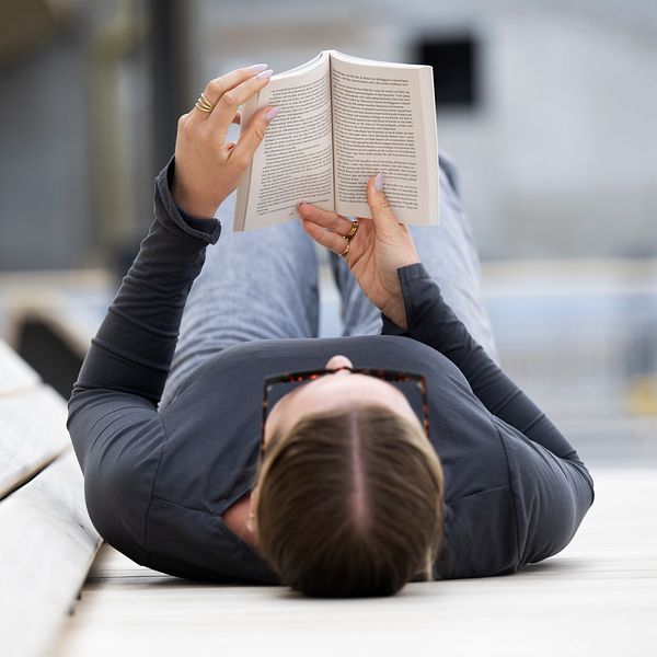 Person ligger på rygg utomhus och läser en bok, kaffemugg står bredvid.