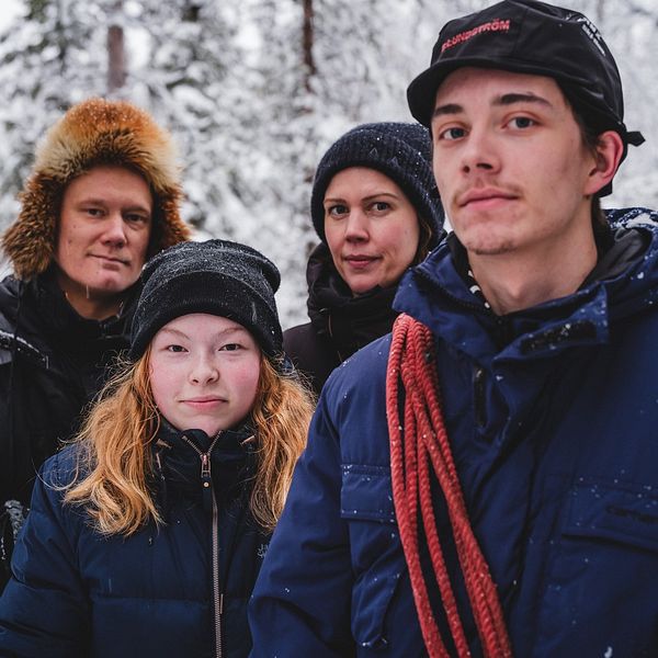 Fyra personer som står utomhus i ett snöigt skogslandskap, klädda i varma vinterkläder. Personen längst till höger bär en mörk blå jacka med en röd rep över axeln och en mörk mössa med skärm. Träden i bakgrunden är täckta av snö.