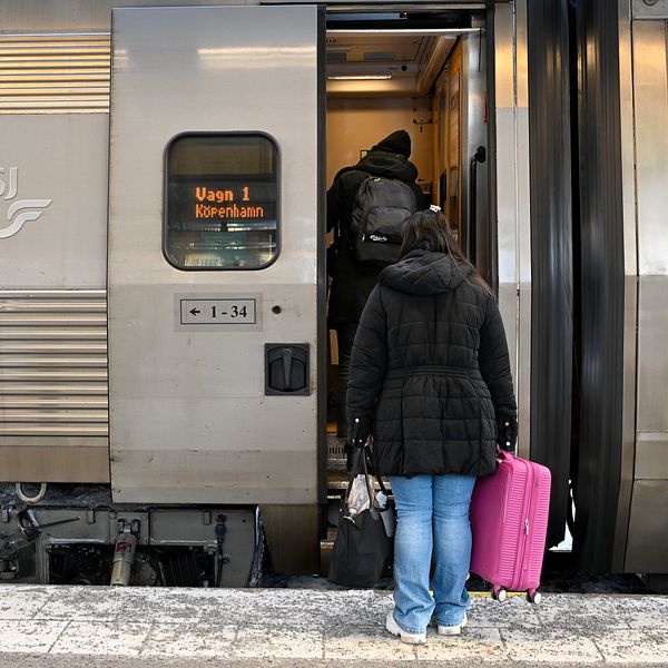En resenär med en rosa resväska går ombord på ett tåg vid en plattform, med tågdörren öppen och en annan passagerare som redan har klivit in. Tåget är märkt med SJ och destinationen Köpenhamn.