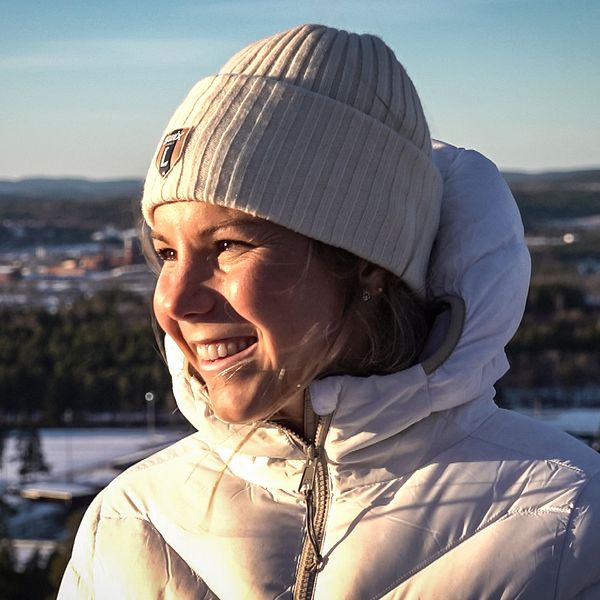 Längdskidåkaren Johanna Hagström iklädd en vit jacka och en ruta med en stickad mössa står vid en utsiktsplats i Falun. Bakgrunden visar en vinterlandskap med snö, träd och en fjärran vy av täckta kullar och en sjö. Dagsljuset skapar en klar och kall atmosfär.