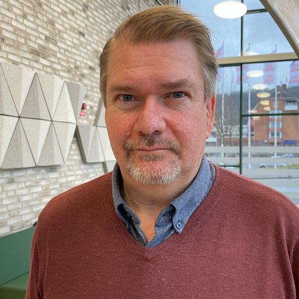 En person står inomhus framför en tegelvägg med geometriska dekorativa paneler, ett modernt mötesrum med en soffa och fönster som visar en stadsbild utanför.