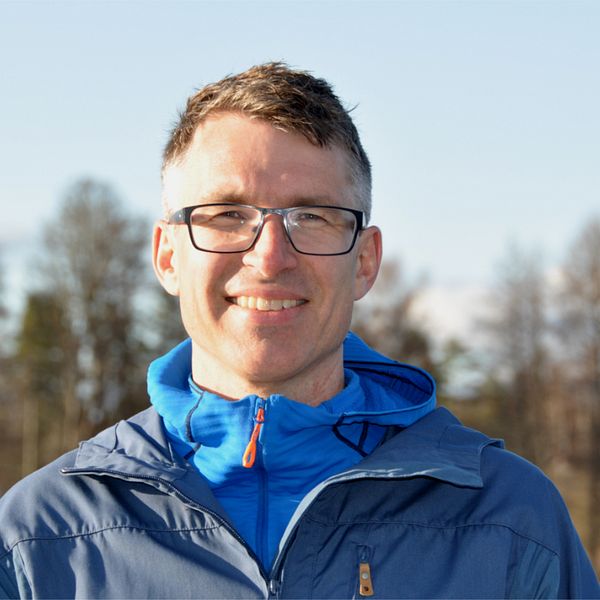 En person i en blå jacka står framför en sjö eller en stor vattensamling, med klart väder och träd i bakgrunden. Det ger en känsla av utomhusaktiviteter och natur.