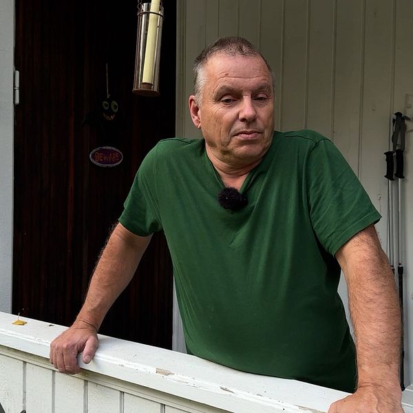 En person står lutad mot en vit träbalustrad på en veranda med en röd trävägg i bakgrunden. Verandan har ett par blommor i en kruka bredvid personen, och det hänger en ljuskrona från taket.