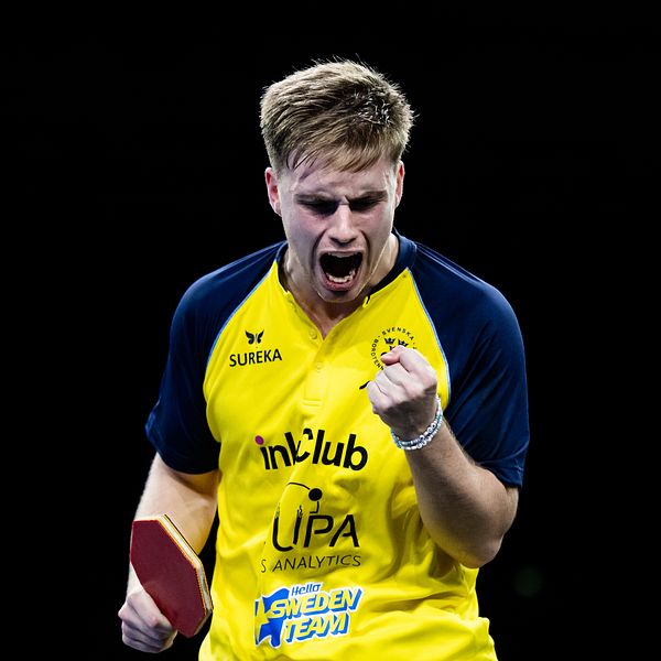 En bordtennisspelare i gul lagdräkt med tryck från sponsorer håller en racket och visar en gest av triumf med sin knutna näve.
