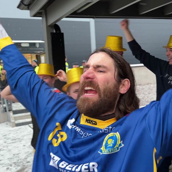 Flera personer firar utomhus, klädda i blå och gula sporttröjor nummer 13 och 10 samt guldhattar, med snö eller hagel på marken. I bakgrunden syns säkerhetspersonal och en byggnad.