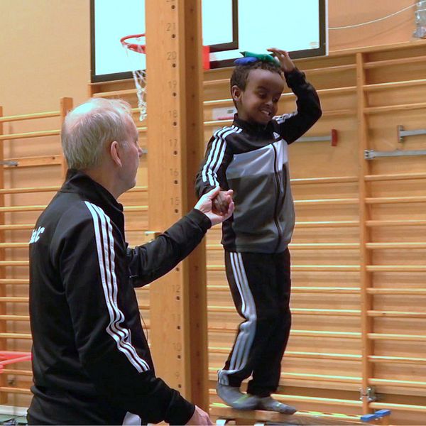 Barn på balansbom instrueras av tränare i gymnastiksal.