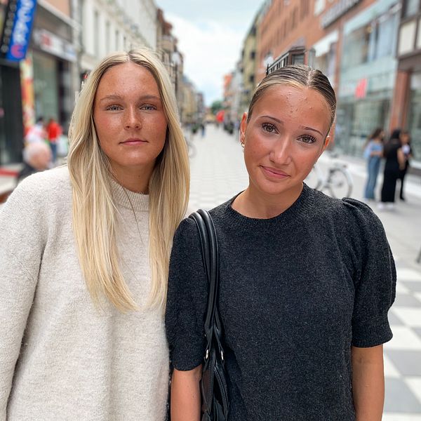 Två tjejer står på en shoppingata och tittar in i kameran.