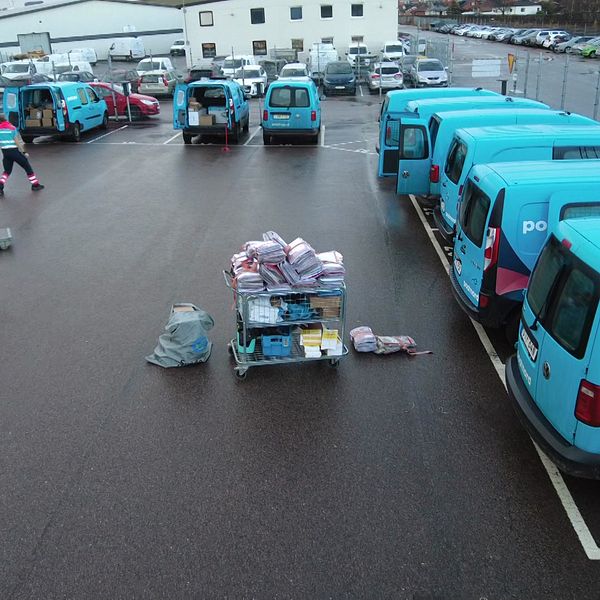 En företagsparkering med flera blå eldrivna transportbilar, där några är öppna och lastade med paket. I förgrunden finns en lastpall med färgglada paket och försändelser, medan två arbetare i reflexvästar förbereder lastning. Området är vått, vilket tyder på nyligen fallande regn.