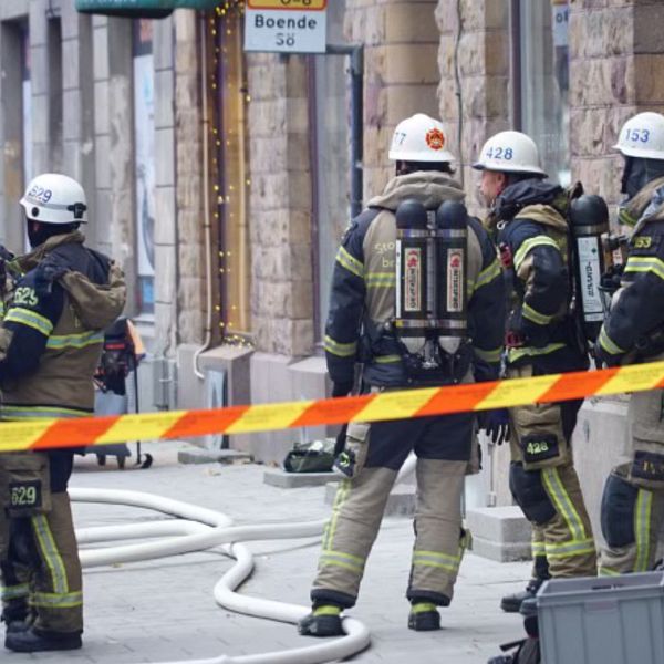 Fyra brandmän i skyddsutrustning står utanför en byggnad på Södermalm i Stockholm, omgivna av brandbilar och brandslangar. De diskuterar situationen medan en av dem håller en släckutrustning. En av brandbilarna syns till vänster, och ett avståndsband markerar avspärrningen runt området.