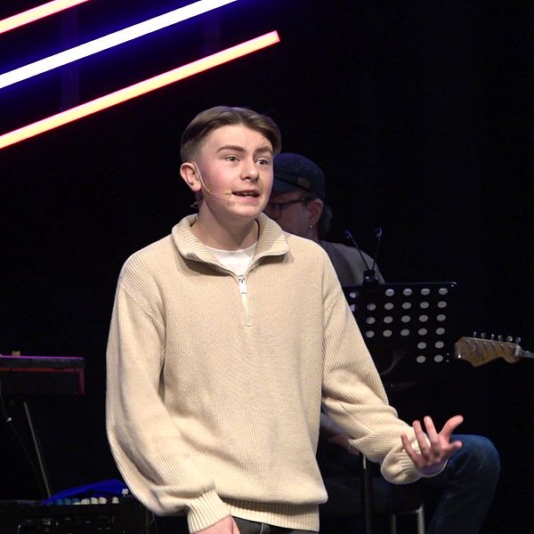 En ung man står på en scen  under en musikalisk framträdande. Bakom honom syns musiker som spelar instrument. Scenens belysning består av färgglada lysrör.