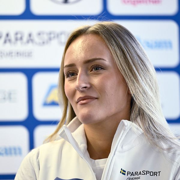 En person iklädd en vit jacka med ”Parasport Sverige” logotyp står framför en bakgrund med olika sponsorer inom idrott.