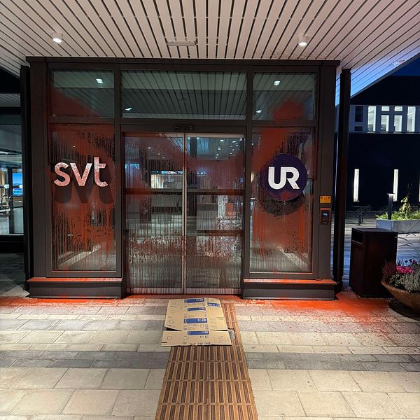 Entrén till en byggnad med logotyperna ”svt” och ”UR” synliga på glasdörrarna. Dörrarna är fläckade med röd färg, vilket ger intryck av klotter eller vandalism. Framför dörrarna ligger en kartong och markeringen på golvet indikerar en gångväg. Belysningen i omgivningen är svag, vilket tyder på att bilden är tagen på natten.