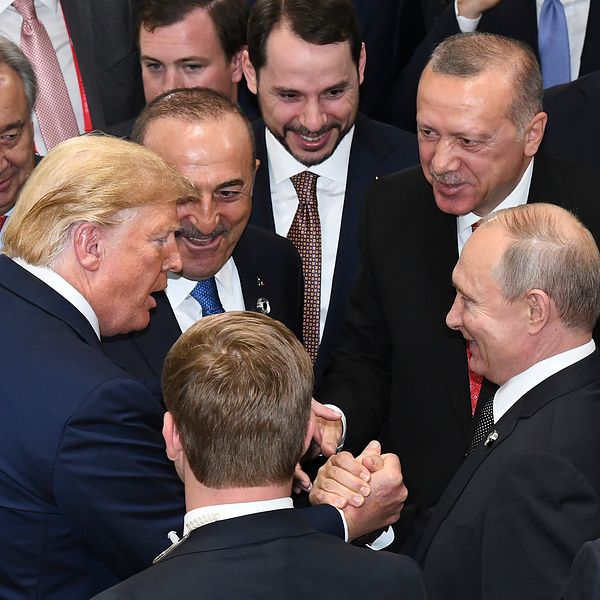 Donald Trump och Vladimir Putin skakar hand och deltar i en konversation bland en grupp män i kostymer.