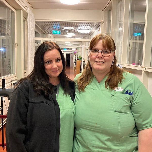 Saana Mänttäri och Anna-Lena Ramsten står i arbetskläder tillsammans framför en ljus korridor. I bakgrunden syns fönster och en rullstol i en vårdmiljö.