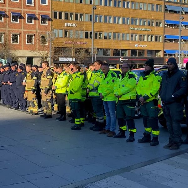 En grupp av räddningspersonal och åskådare står uppställda vid ett torg under en ceremoni eller demonstration framför två större byggnader.