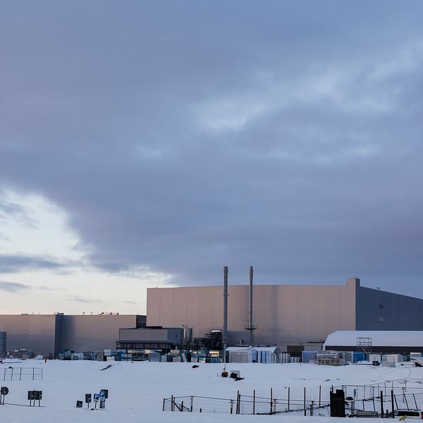 Industriområde med stora byggnader täckta av snö under en molnig himmel. Den främre byggnaden har två rör som sträcker sig mot himlen, medan en mindre byggnad syns till höger. Landskapet präglas av en vintrig miljö och få aktiviteter syns runt området.
