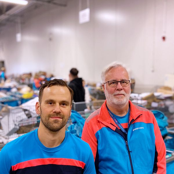 Två personer står i en lagerlokal omgiven av handkorgar och emballage. Den ena personen bär en blå t-shirt med röd detalj, medan den andra har på sig en färgglad jacka. I bakgrunden syns en stor mängd varor och korgar organiserade i rader. En väggklocka hänger på väggen, vilket visar att det är dagsljus.
