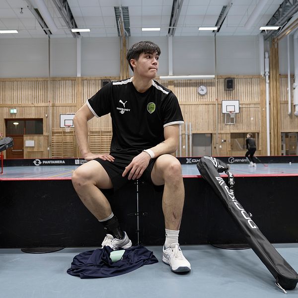 Två innebandyspelare sitter på en bänk i en sporthall med klubbor bredvid sig.