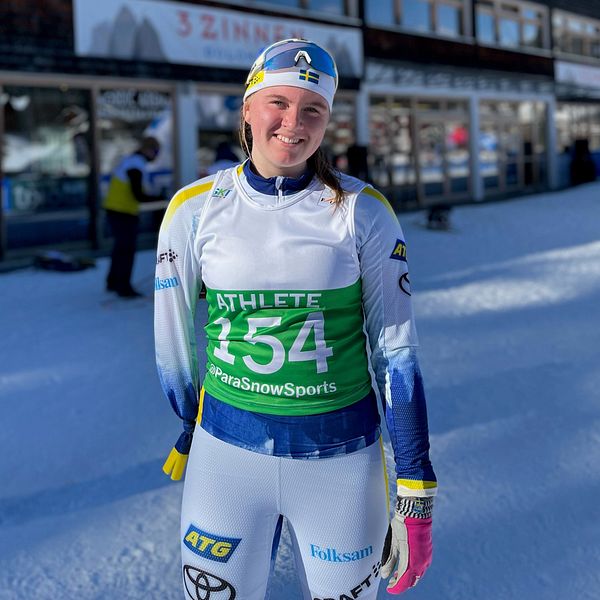 En skidåkare klädd i tävlingsdräkt står framför Toblach Nordic Arena, med klar himmel och snö runt omkring. Skidåkaren har startnummer 154 på en grön väst. Bakgrunden visar arenans mörka fasad och fönster.