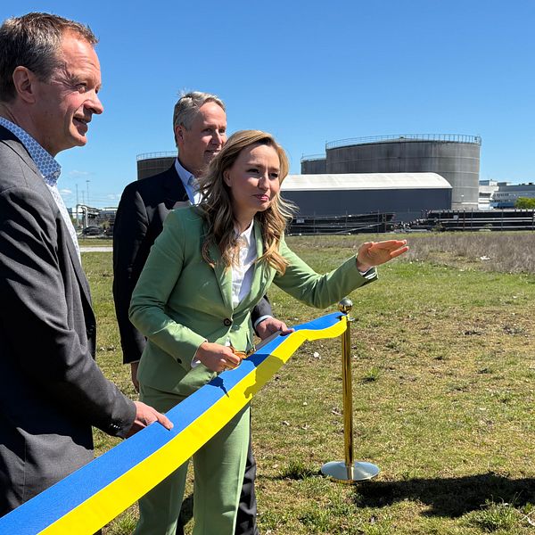 Ebba Busch (KD) i grön kostym klipper blågul band utomhus vid industriområde, tre män närvarande.