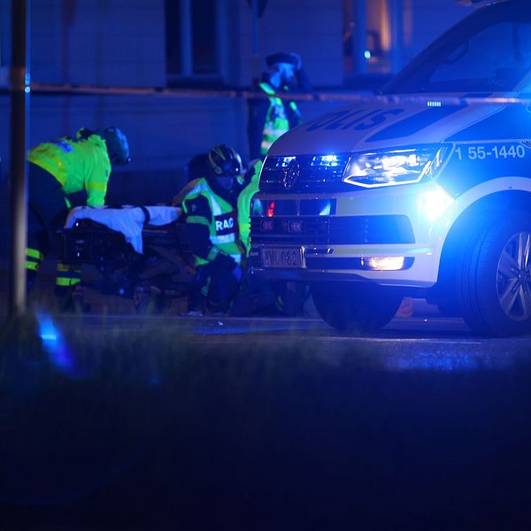 En polisbuss med blåljusen på står bakom en polisavspärrning, bredvid arbetar ambulanspersonal.