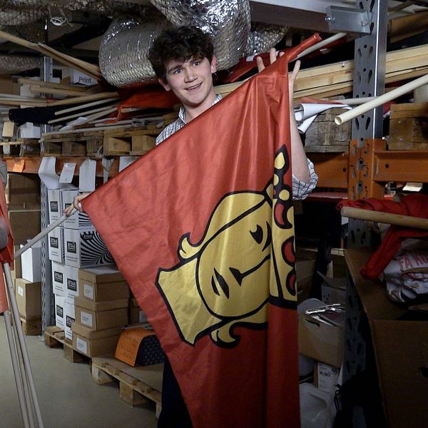 Två personer står i ett lager och håller i en röd flagga med ett gult ansikte.