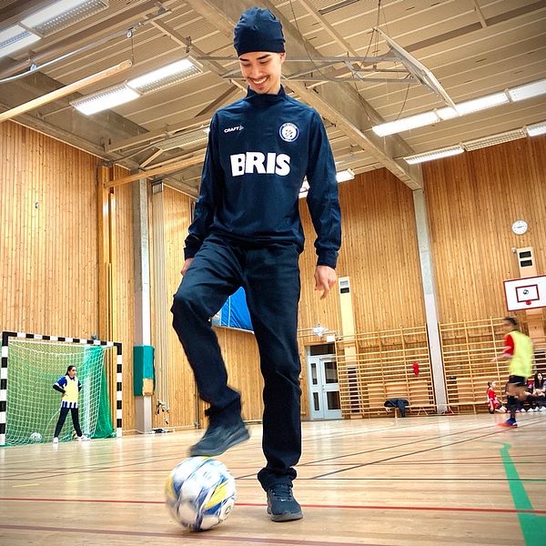 En person står i en sporthall och tränar fotboll, med ena foten som stöttar en fotboll på golvet. I bakgrunden syns en grön målvaktsbur och flera spelare som övar. Väggarna är av trä och rummet har god belysning.