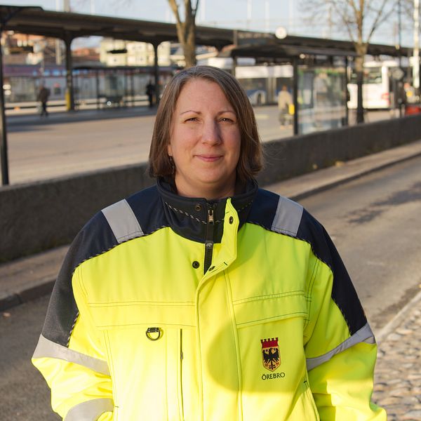 En person klädd i en neonfärgad arbetsjacka med reflexdetaljer står vid en busshållplats. I bakgrunden syns en stadsmiljö med bussar och andra människor som väntar.