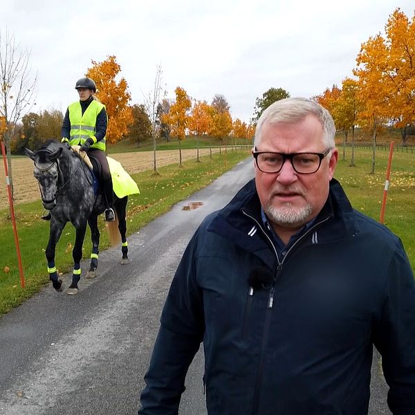 Man i mörk jacka på en väg. Bakom till vänster i bild en ryttare med reflexväst på en häst, som också har reflexer.