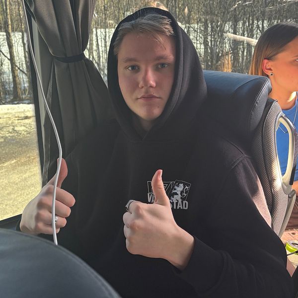 Två personer på buss, en ger tummen upp. Skog ses genom fönstret.
