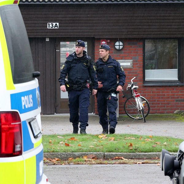 Två poliser i uniform går nära en byggnad. I bakgrunden syns en cykel, och i förgrunden står en polisbil.
