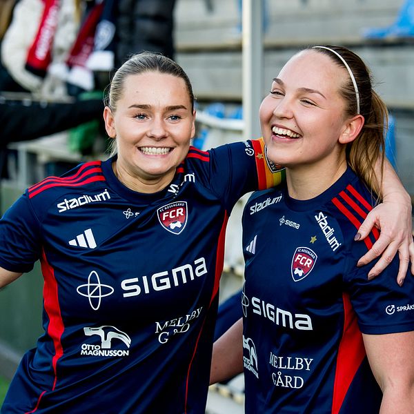 Två fotbollsspelare i blå matchställ firar tillsammans på en fotbollsarena.