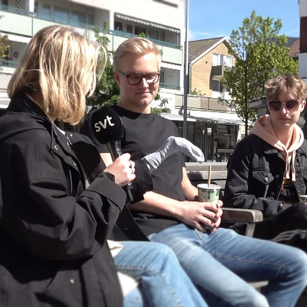 Reporter intervjuar två personer på bänk utomhus i stadsmiljö.
