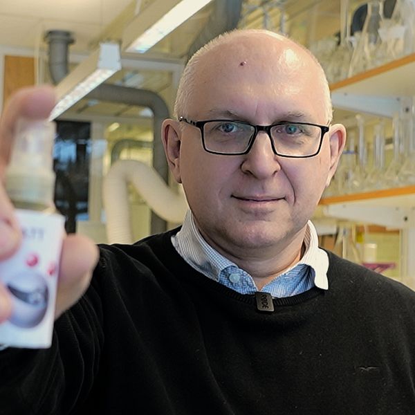 Person i labbrock håller en liten flaska i ett laboratorium fullt med utrustning och glasbehållare i hyllor.