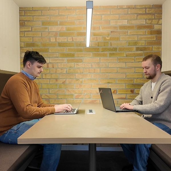 Två personer sitter vid ett bord i en avskild arbetsmiljö, båda arbetar på laptops.