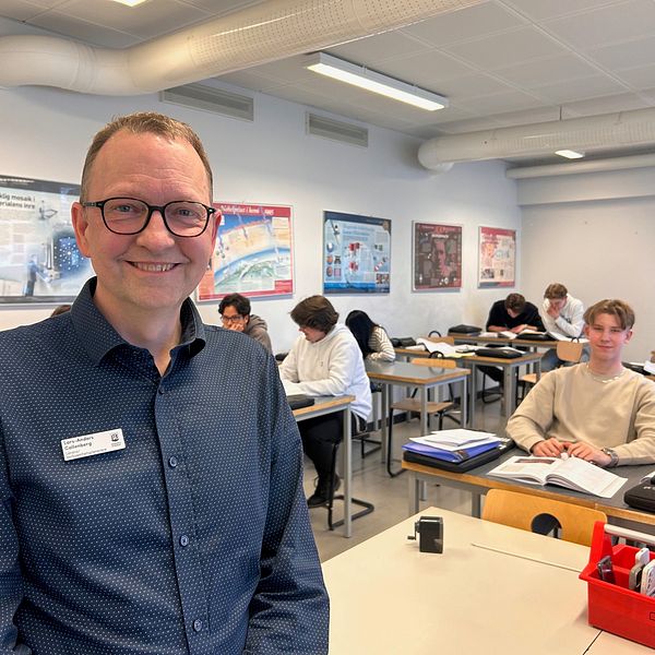 Klassrum med elever vid skrivbord och en lärare i förgrunden. han ler mot kameran.