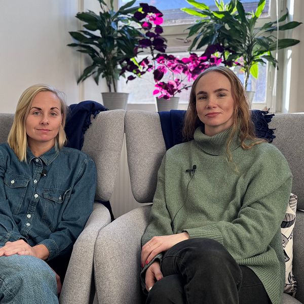 Två personer sitter i bekväma fåtöljer i ett ljust rum med växter och blommor i bakgrunden. Rummet har en avslappnad atmosfär, och det syns en whiteboard på väggen. Den ena personen bär en jeansjacka och den andra en grön tröja.