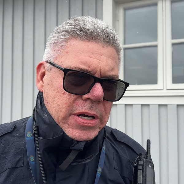 Person med radiokommunikationsutrustning framför grått hus med vita fönster.