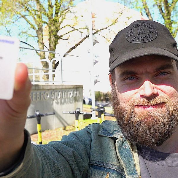 En skäggig man med svart keps och jeansjack håller upp ett körkort utomhus framför en byggnad och träd.