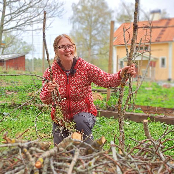 Kvinna står med kvistar och grenar i hand bakom en rishög framför sitt hus i jakt med att välkomna insekter