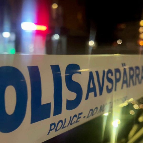 En närbild av ett avspärrat område med en polisavspärrningsbanderoll som visar texten ”POLIS AVSPÄRRAT” och ”POLICE DO NOT CROSS”. I bakgrunden syns suddiga ljus från polisfordon.