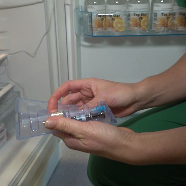 Till vänster är ett kylskåp, till höger i bild en persons händer syns hålla i vaccinsprutor. Personen har gröna kläder.